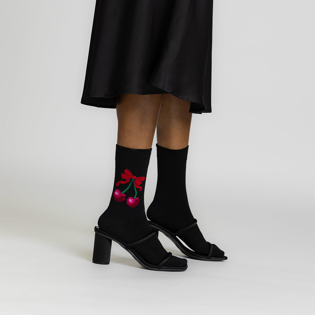 A person wearing black mid-heel sandals and black crew socks with a red cherry and bow graphic, shown from the knees down against a light gray studio background.