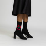 A person wearing black mid-heel sandals and black crew socks with a red cherry and bow graphic, shown from the knees down against a light gray studio background. thumbnail
