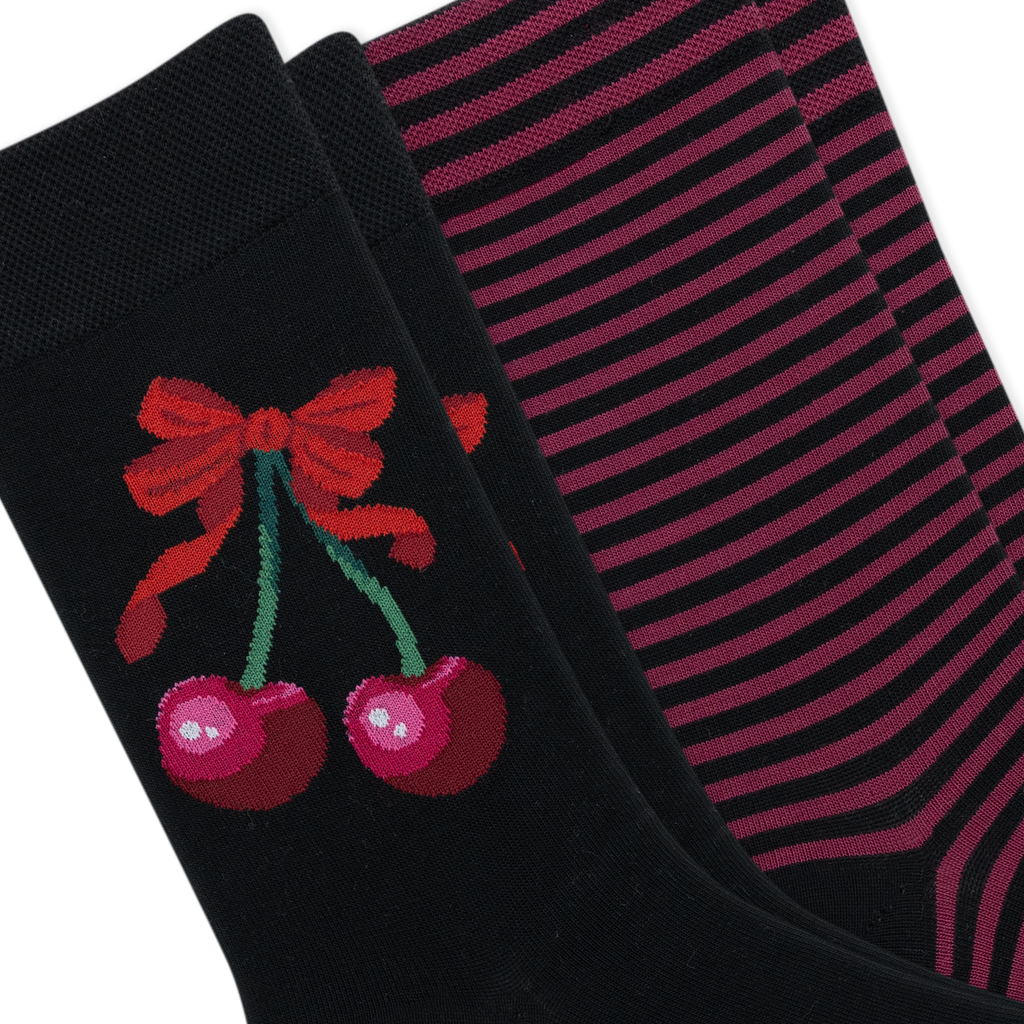 Two pairs of black crew socks arranged diagonally. The front sock features a red bow and two pink cherries with green stems, while the back pair has thin horizontal pink stripes.