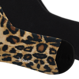 A close-up of a pair of crew socks, with one sock showing a leopard print pattern in beige, brown, and black, and the other sock showing a solid black knit. The brand name 'k.bell' is on the bottom of the foot.