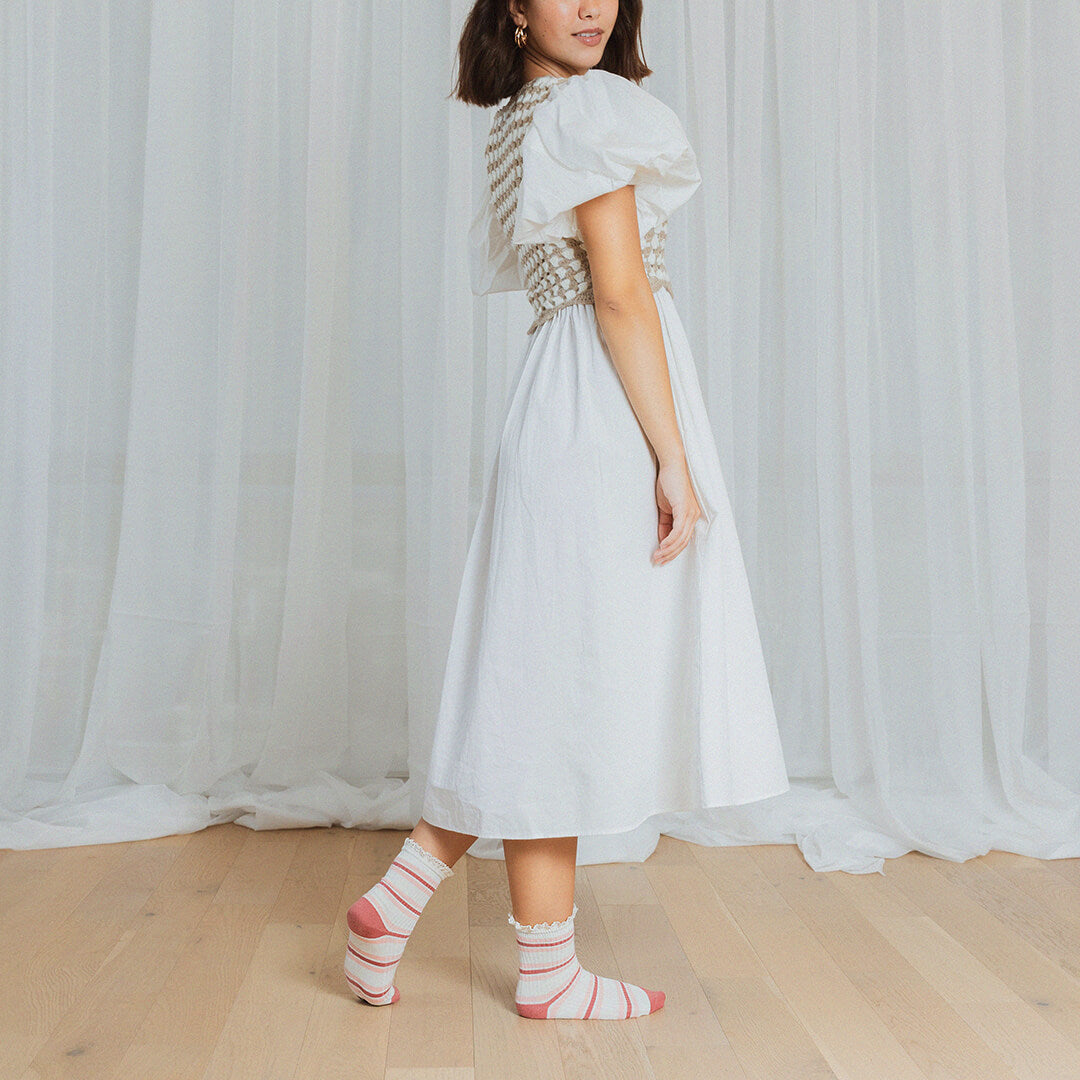 Woman wearing a white dress with a patterned pink and white striped socks standing in front of white curtains.