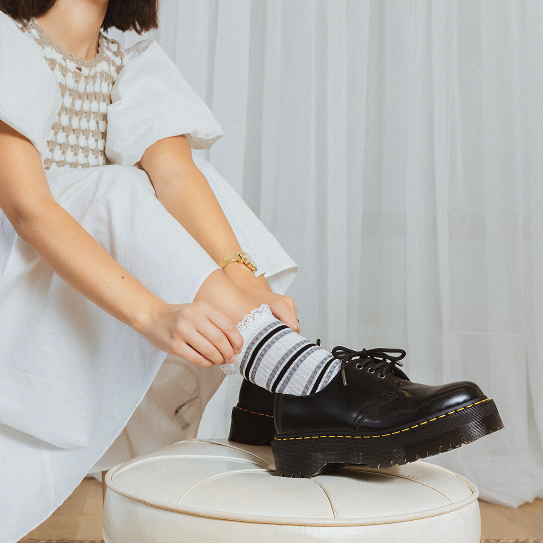 Woman wearing a white dress with a patterned black and white striped socks sitting in front of white curtains.