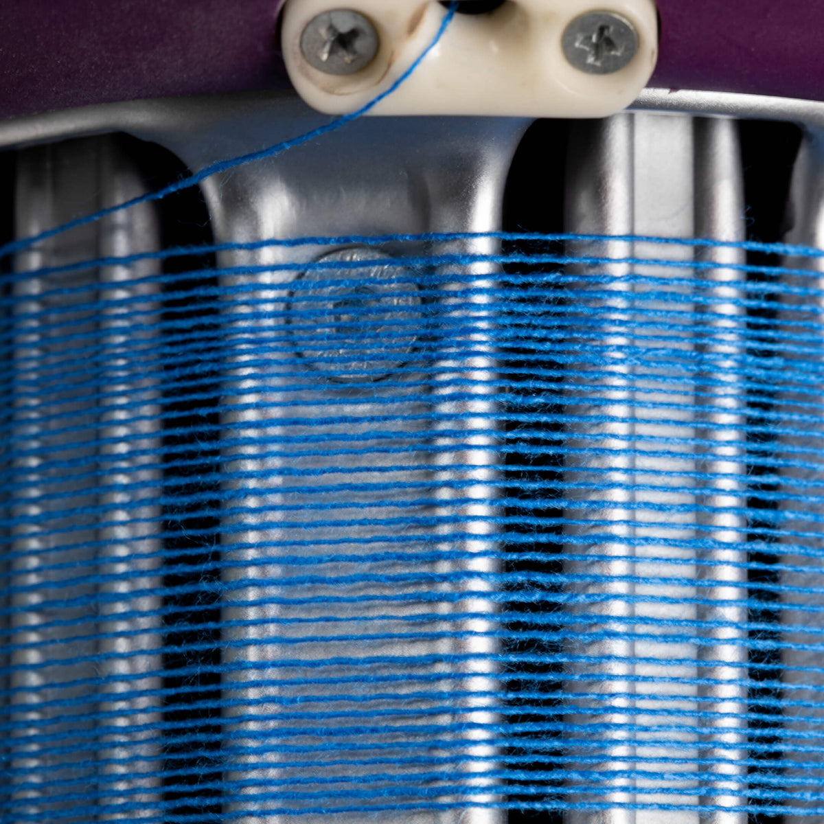 A close-up of bright blue yarn wound tightly around a metallic spool, with part of a purple fabric or material visible at the top and a cream plastic guide holding the thread.
