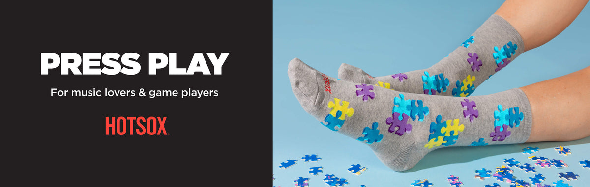 Gray crew socks with colorful jigsaw puzzle pattern, worn on feet against a light blue background with puzzle pieces scattered, alongside promotional text 'PRESS PLAY For music lovers & game players' and the HOTSOX brand.