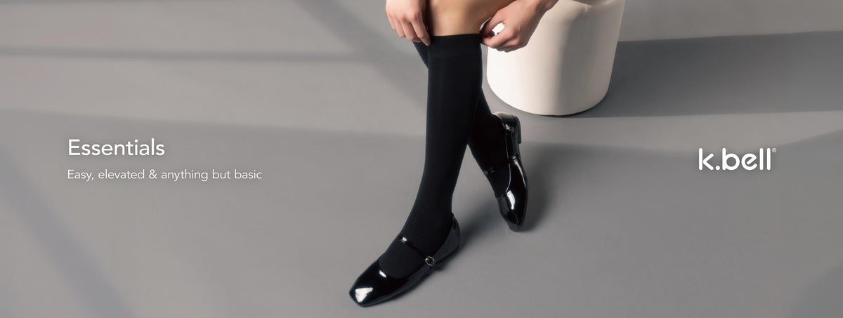 A model wearing black knee-high socks and black pointed-toe flats with silver embellishments, standing on a gray floor next to a white cylindrical stool. The text on the left reads 'Essentials – Easy, elevated & anything but basic' and the 'k.bell' logo