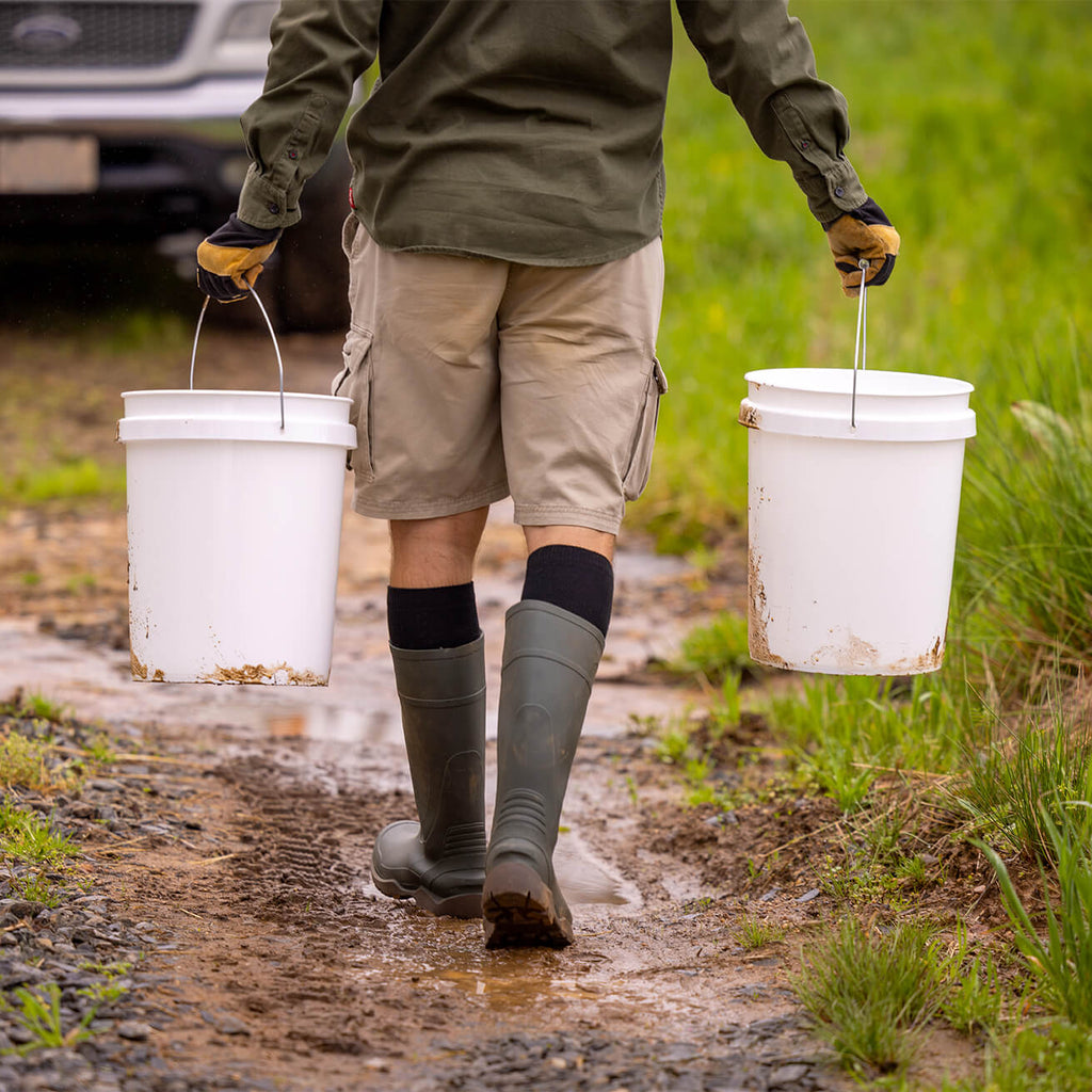 Person walking on a muddy path carrying two white buckets.