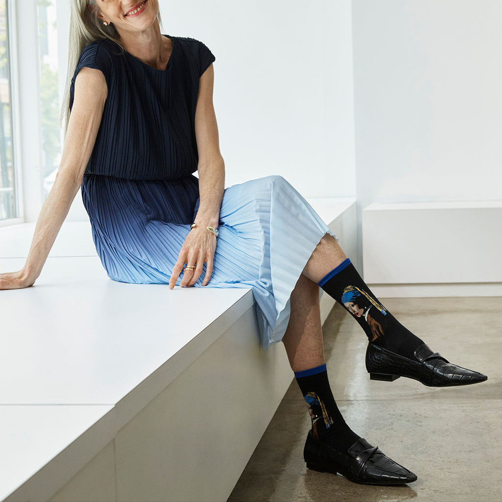 Woman sitting on a white bench wearing black dress shoes and socks with a design.