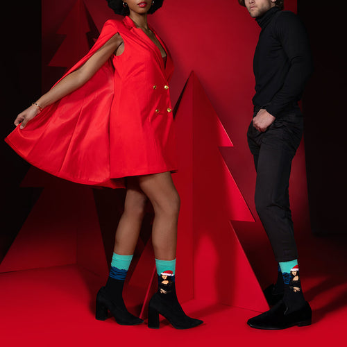 Person wearing a red dress with a cape and another person in black attire on a red background wearing Santa-themed Mona Lisa socks