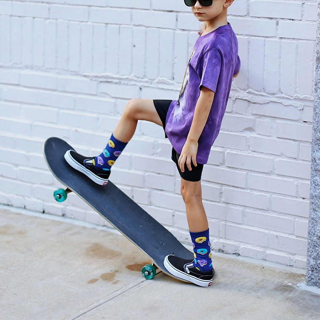 Child in purple shirt and black shorts standing on a skateboard against a white brick wall.