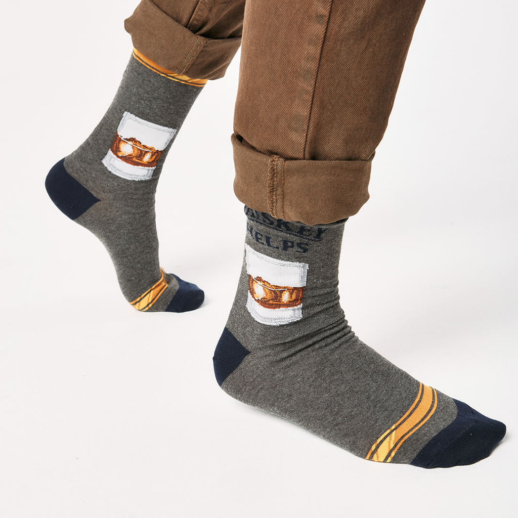 Gray socks with a whiskey glass graphic and text that says "Whiskey Helps"  worn with brown pants on a white background