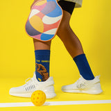 Person wearing colorful socks with 'The Dinker' designs, white sneakers, and a pickleball paddle on a yellow background.