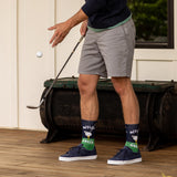 Person wearing socks with golf-themed designs, standing on a wooden deck with a golf club and ball. thumbnail