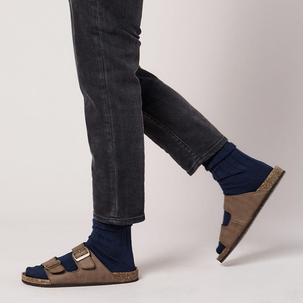 Brown sandals worn with dark jeans and navy socks on a light gray background