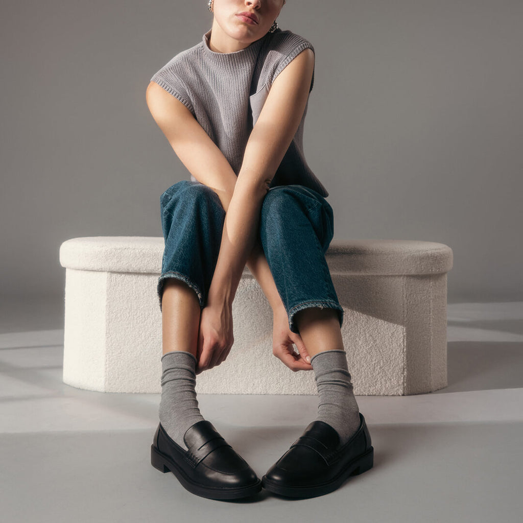 Person sitting on a textured ottoman wearing a sleeveless top, jeans, and black shoes with gray socks.