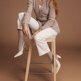 Woman sitting on a stool wearing a beige cardigan and socks, white pants, and high-heeled shoes against a brown background