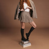Person wearing a plaid skirt and jacket with brown knee-high socks and shoes on a brown background