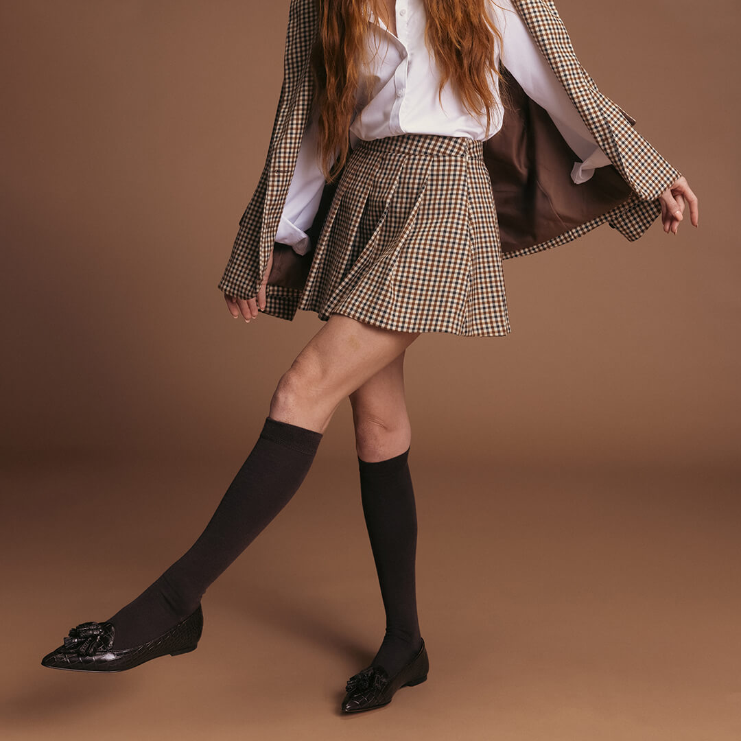 Person wearing a plaid jacket, white shirt, brown skirt, brown shoes, and brown socks on a brown background