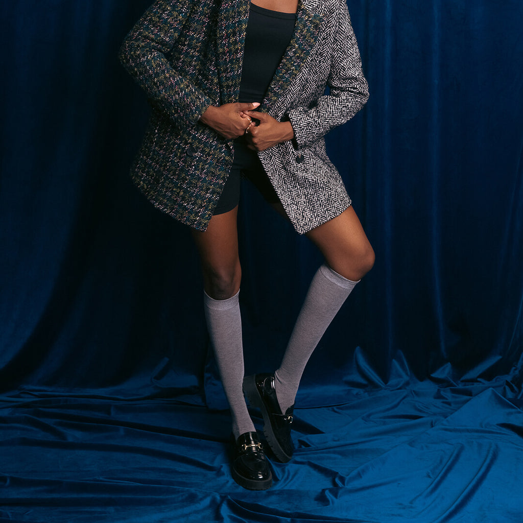 Person wearing a patterned blazer, black top, knee-high grey socks, and black shoes on a blue background