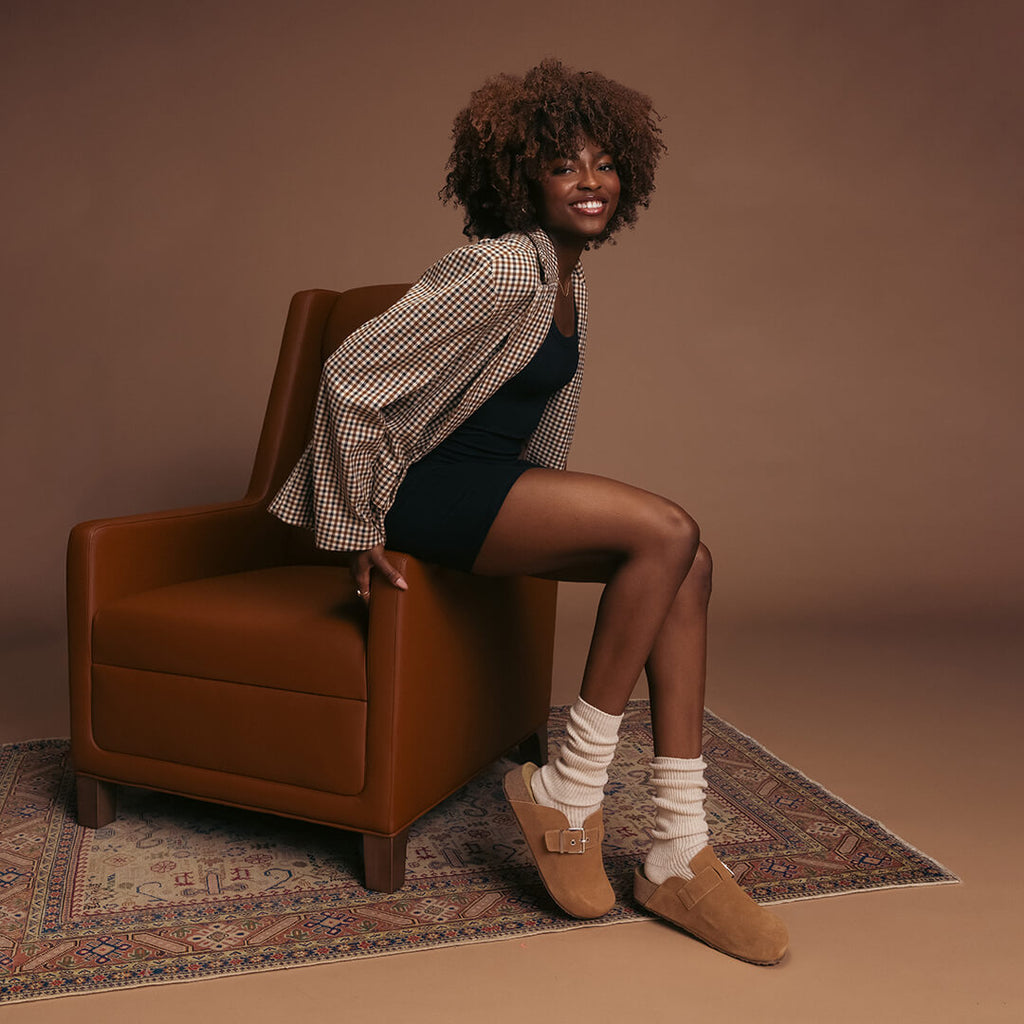 Woman sitting on a brown armchair wearing a checkered shirt, black dress, beige slouch socks and brown shoes on a brown background.