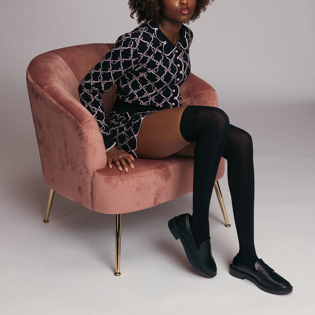 Person sitting on a pink chair wearing a patterned black and white outfit with black thigh-high socks and shoes.