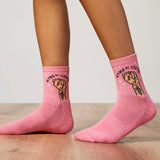 Pink socks with fist design and 'Women Unite' text worn by a person on a wooden floor. thumbnail