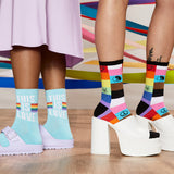 Two pairs of feet wearing colorful socks with messages and designs, standing on a wooden floor. thumbnail