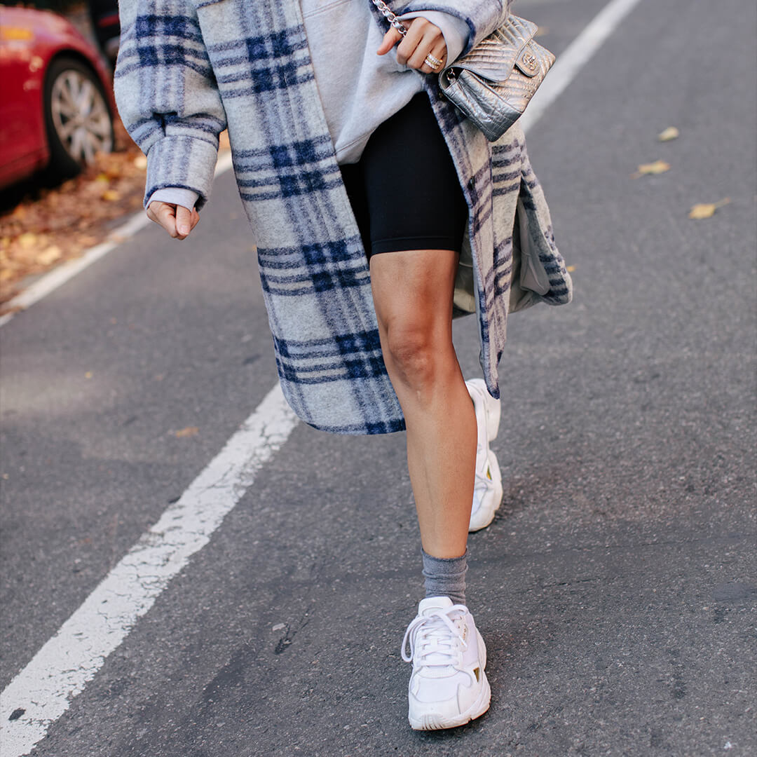 Person wearing a plaid coat, black shorts, white sneakers, and gray roll top socks on a street.