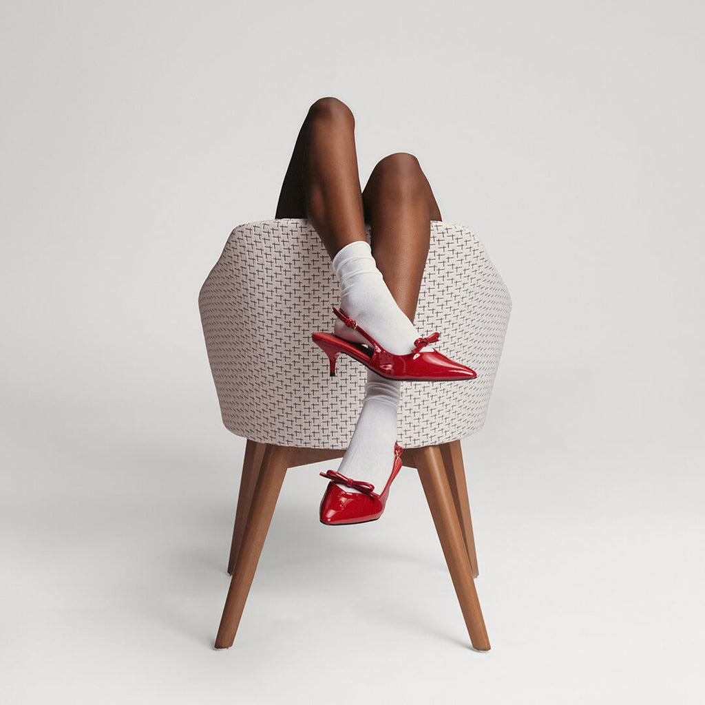 Person wearing red high-heeled shoes and white socks sitting on a woven stool against a plain background